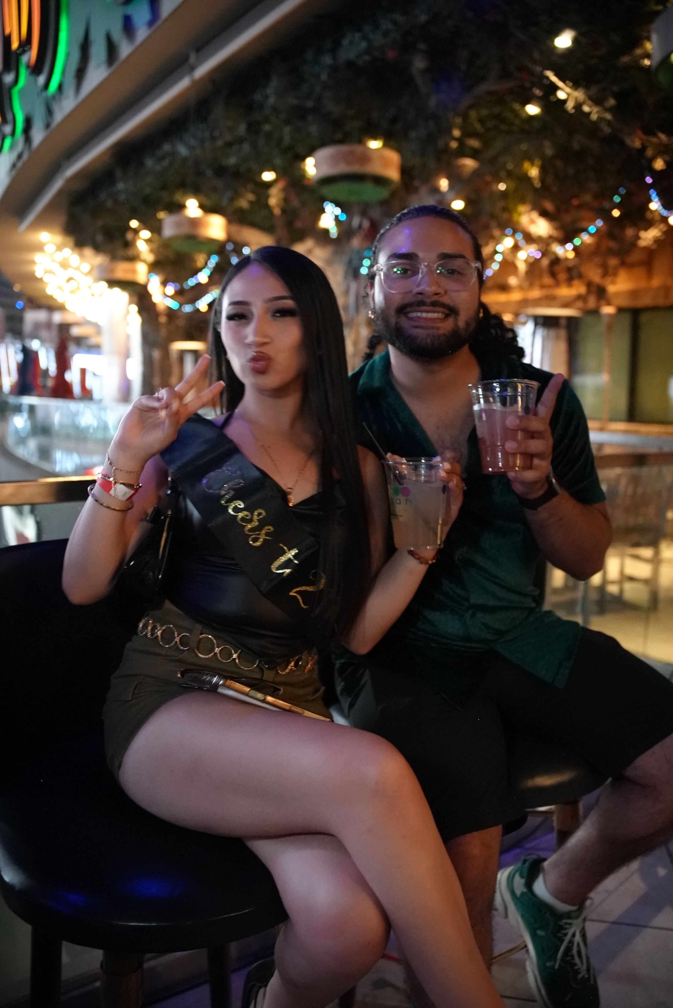 Two friends posing with cocktails on bar stools during a night out; woman wearing a celebratory sash flashes a peace sign under colorful string lights and tropical decor in a lively bar interior.
