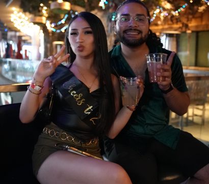 Two friends posing with cocktails on bar stools during a night out; woman wearing a celebratory sash flashes a peace sign under colorful string lights and tropical decor in a lively bar interior.