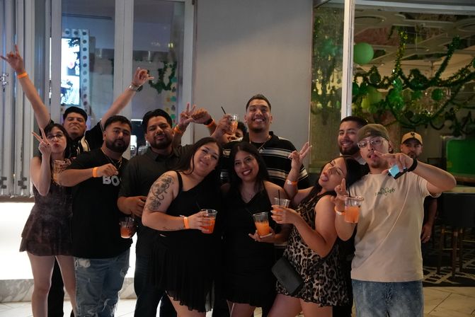 Group of friends at an indoor bar lounge on a night out, smiling and posing with orange cocktails and event wristbands, festive green decorations in the background