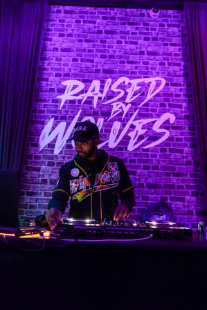 DJ mixing on turntables in a vibrant purple LED-lit nightclub, performing in front of a brick wall mural that reads "RAISED BY WOLVES".