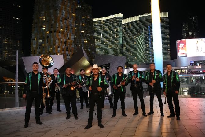 Energetic Mexican brass band in matching green-sequin jackets and a cowboy-hat frontman posing with trombones, sousaphone, trumpet and clarinet on a Las Vegas Strip plaza at night with lit skyscrapers.