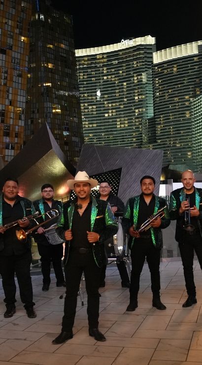 Energetic Mexican brass band in matching green-sequin jackets and a cowboy-hat frontman posing with trombones, sousaphone, trumpet and clarinet on a Las Vegas Strip plaza at night with lit skyscrapers.