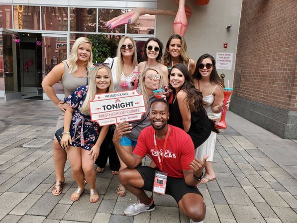 Smiling group of friends with an event guide posing on a downtown plaza under a large pink flamingo sculpture, holding colorful souvenir drink cups and a red-and-white sign reading “TONIGHT” for a nightlife pub crawl.