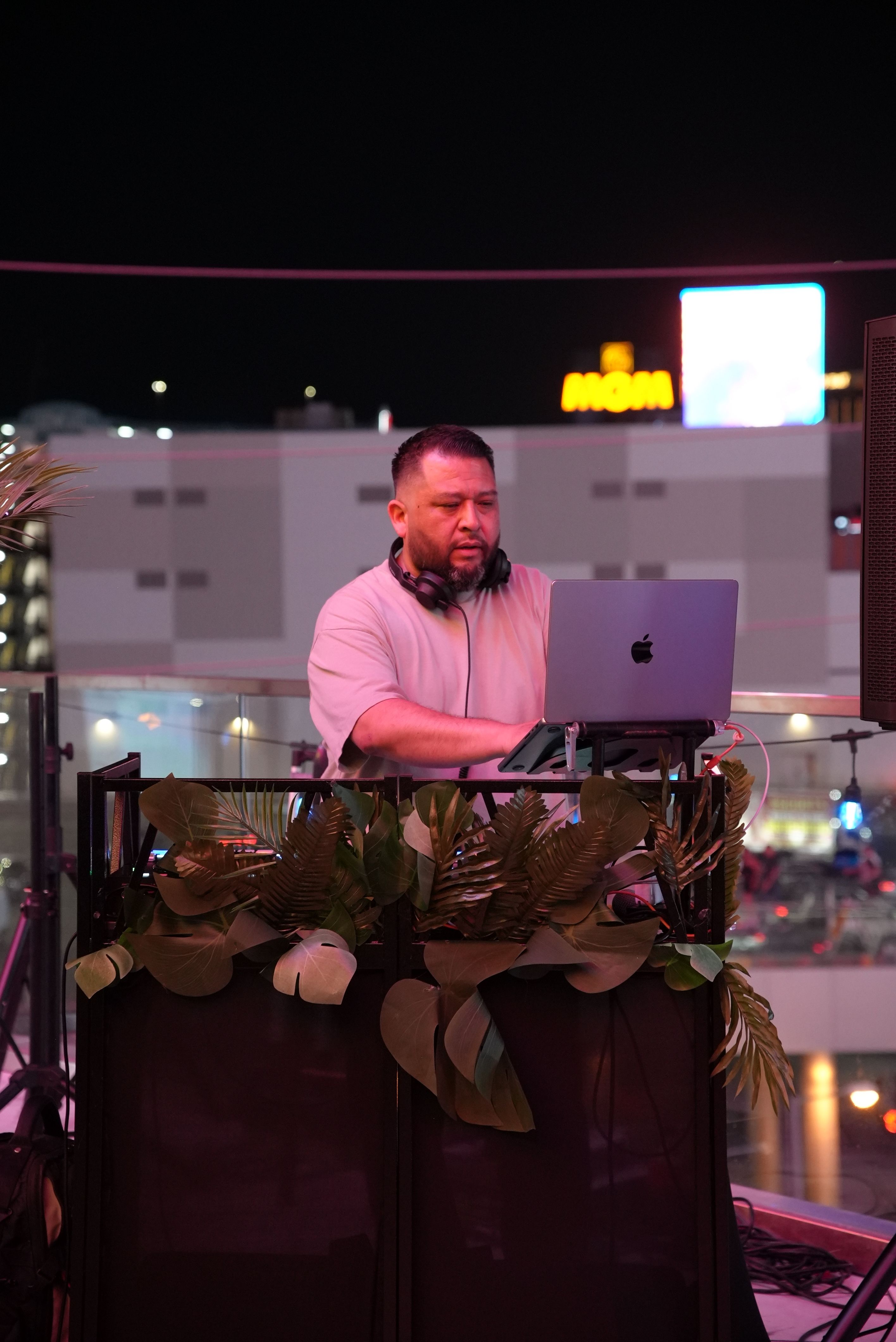 Rooftop DJ spinning a nighttime city set on a laptop behind a tropical plant–decorated booth, colorful neon lights glowing in the background