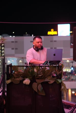 Rooftop DJ spinning a nighttime city set on a laptop behind a tropical plant–decorated booth, colorful neon lights glowing in the background