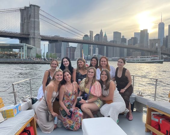 Smiling friends enjoying a sunset boat cruise on the East River with the Brooklyn Bridge and Manhattan skyline in New York City.