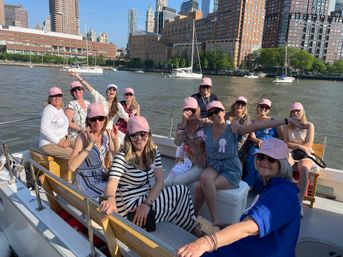 Cheerful group of women in matching pink caps enjoying a sunny boat cruise with sailboats on the Hudson River and New York City skyline in the background