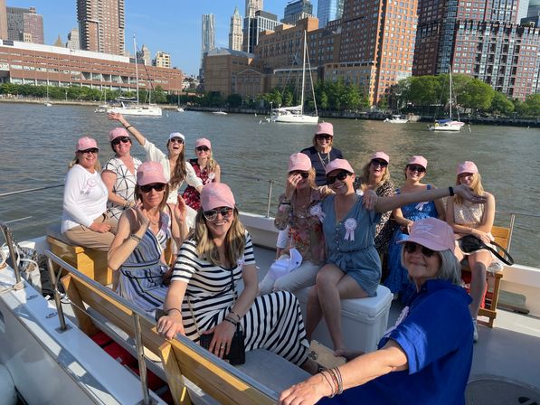 Cheerful group of women in matching pink caps enjoying a sunny boat cruise with sailboats on the Hudson River and New York City skyline in the background