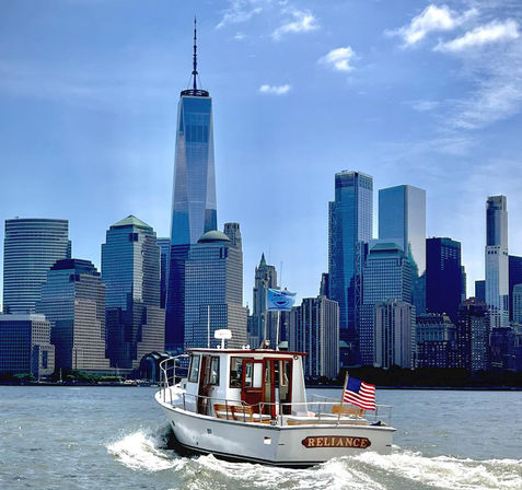 White cabin motorboat flying an American flag cuts through New York Harbor with the Lower Manhattan skyline and One World Trade Center towering under a bright blue sky.