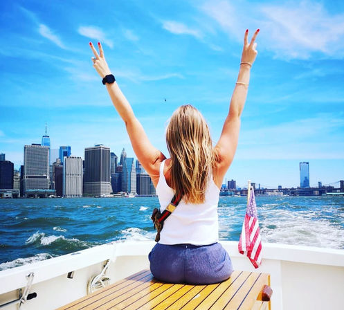 Woman sitting on a boat with arms raised in peace signs, facing the Lower Manhattan skyline and Brooklyn Bridge over choppy blue water, American flag on the stern under a bright sunny sky