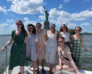 Seven friends in summer dresses wearing pink heart-shaped sunglasses posing on a boat in New York Harbor with the Statue of Liberty and a bright blue, cloud-dotted sky behind them.