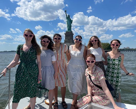 Seven friends in summer dresses wearing pink heart-shaped sunglasses posing on a boat in New York Harbor with the Statue of Liberty and a bright blue, cloud-dotted sky behind them.