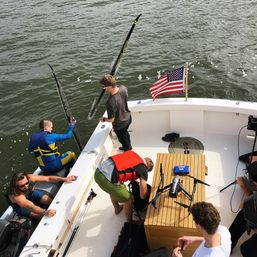 Overhead shot of people on a boat wearing life vests, prepping water-skis and a drone on the teak deck beside an American flag over choppy water.