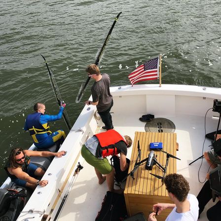 Overhead shot of people on a boat wearing life vests, prepping water-skis and a drone on the teak deck beside an American flag over choppy water.