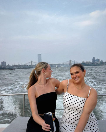 Two friends on a boat ride on the East River, smiling in summer outfits with the Manhattan skyline and Brooklyn Bridge in the background
