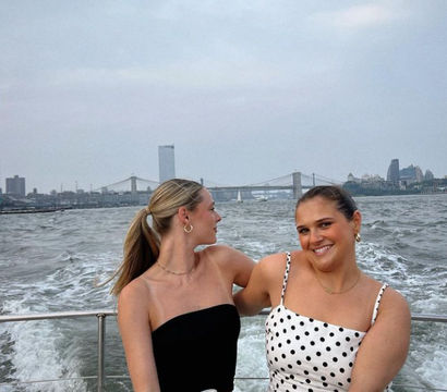 Two friends on a boat ride on the East River, smiling in summer outfits with the Manhattan skyline and Brooklyn Bridge in the background