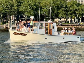 White motorboat cruising along a tree-lined urban waterfront with passengers on deck enjoying a sunny afternoon.