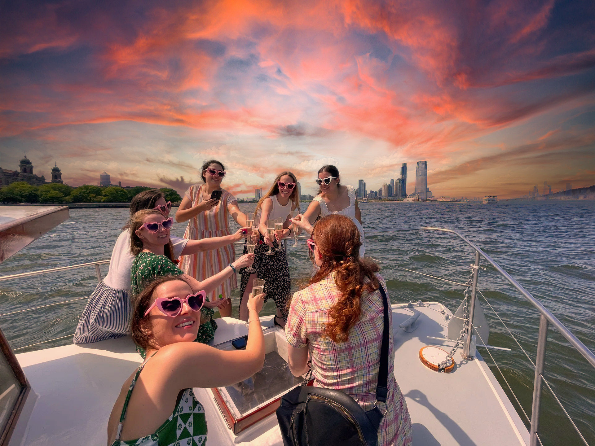 Group of friends on a boat toasting with champagne and wearing heart-shaped sunglasses, vibrant pink-orange sunset over a distant city skyline and waterfront.