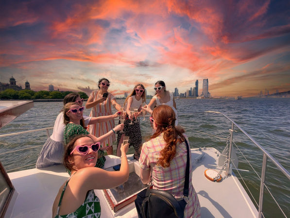 Group of friends on a boat toasting with champagne and wearing heart-shaped sunglasses, vibrant pink-orange sunset over a distant city skyline and waterfront.