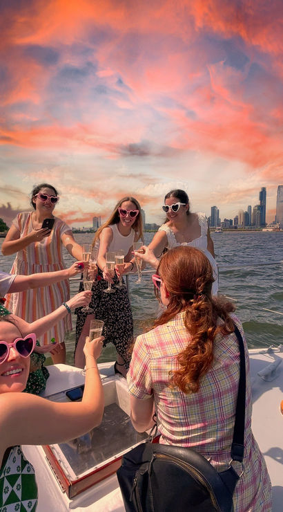 Group of friends on a boat toasting with champagne and wearing heart-shaped sunglasses, vibrant pink-orange sunset over a distant city skyline and waterfront.