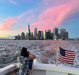 Person on a boat photographing New York City’s Lower Manhattan skyline at sunset — American flag on the stern, choppy harbor water and vivid pink-blue sky with One World Trade Center visible.