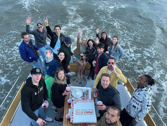 Overhead view of a happy group of friends waving on a boat party at sea, surrounded by wake, pizza boxes and red cups.