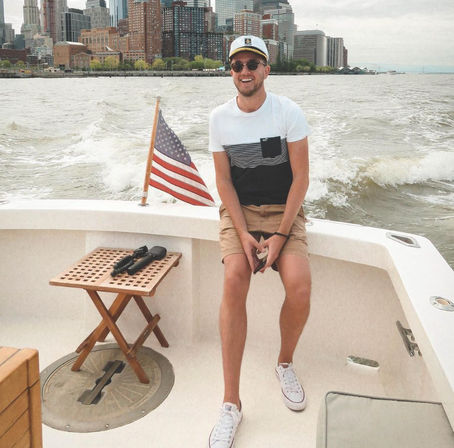 Smiling person in a captain’s hat and sunglasses sitting on a boat with an American flag at the stern, urban skyline and choppy harbor waters in the background.