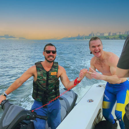 Two men laughing during a jet-ski outing at sunset — one in a camo life vest and sunglasses, the other shirtless in blue-and-yellow swim leggings, clasping hands on a boat with a wake trailing and an urban waterfront skyline in the distance.