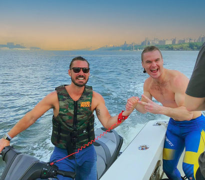 Two men laughing during a jet-ski outing at sunset — one in a camo life vest and sunglasses, the other shirtless in blue-and-yellow swim leggings, clasping hands on a boat with a wake trailing and an urban waterfront skyline in the distance.