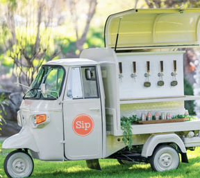 White vintage three-wheeled mobile drink cart with five taps and canned beverages, trimmed with greenery and parked on grass in a sunlit garden setting