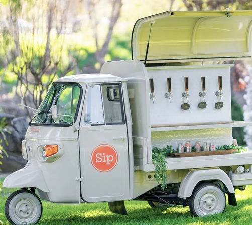 White vintage three-wheeled mobile drink cart with five taps and canned beverages, trimmed with greenery and parked on grass in a sunlit garden setting