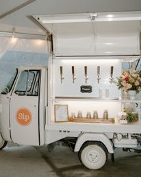 Chic white mobile prosecco bar in an event tent with four taps, backlit hex-tile backsplash, stacked cups on a tray, framed menu and a blush floral arrangement.