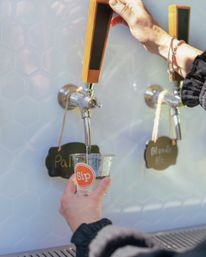Hand pulling a wooden draft tap pouring pale ale into a clear tasting cup at a craft taproom