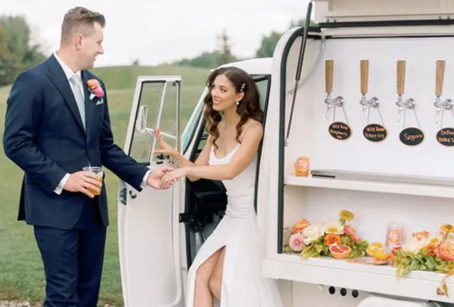 Bride in a white slit wedding dress stepping from a vintage white mobile bar van, taking the hand of her groom in a navy suit holding a drink, outdoor wedding with floral decor and wooden beer taps