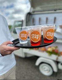 Server holding a tray of plastic cups labeled "Sip" filled with pink and clear drinks in front of a white mobile beverage cart at an outdoor event