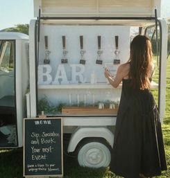 Mobile outdoor bar trailer on a sunny lawn with draft taps, oversized 'BAR' letters, a chalkboard menu advertising event bookings, and a woman in a black dress placing an order.