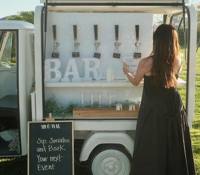 Mobile outdoor bar trailer on a sunny lawn with draft taps, oversized 'BAR' letters, a chalkboard menu advertising event bookings, and a woman in a black dress placing an order.