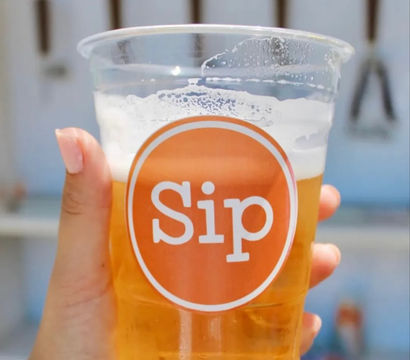 Hand holding a clear plastic cup of golden beer with a frothy head and orange circular logo, blurred outdoor event background