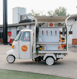 White three-wheeled mobile drink cart parked in an urban lot, open service window with five taps, stacked cups, citrus and floral arrangements, and an orange circular logo on the door.