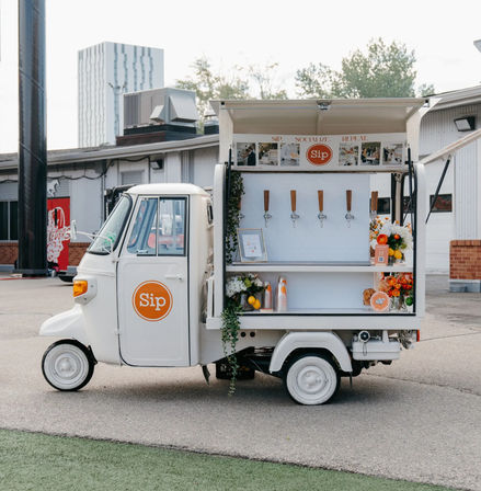 White three-wheeled mobile drink cart parked in an urban lot, open service window with five taps, stacked cups, citrus and floral arrangements, and an orange circular logo on the door.