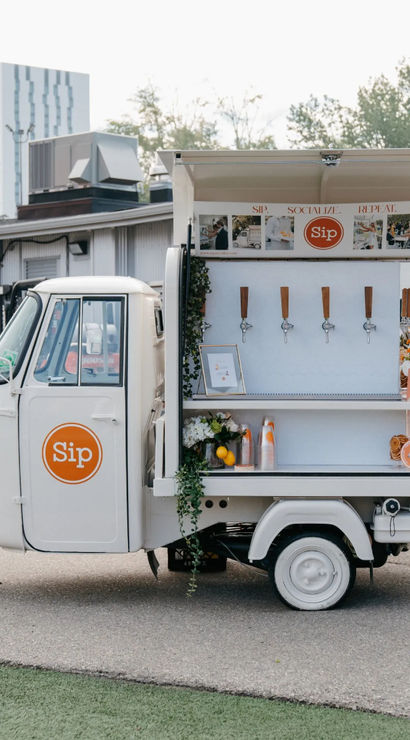 White three-wheeled mobile drink cart parked in an urban lot, open service window with five taps, stacked cups, citrus and floral arrangements, and an orange circular logo on the door.