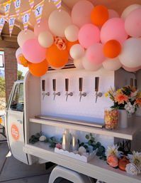 Festive outdoor mobile drink truck with six taps, pink, peach and orange balloon garland, floral arrangements, stacked plastic cups and a jar of candy.