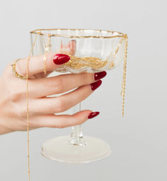 Close-up jewelry display: hand with glossy red manicure and gold rings holds a glass coupe filled with delicate gold chains and necklaces against a light gray background.