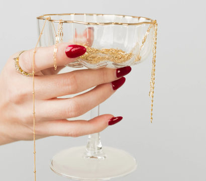Close-up jewelry display: hand with glossy red manicure and gold rings holds a glass coupe filled with delicate gold chains and necklaces against a light gray background.