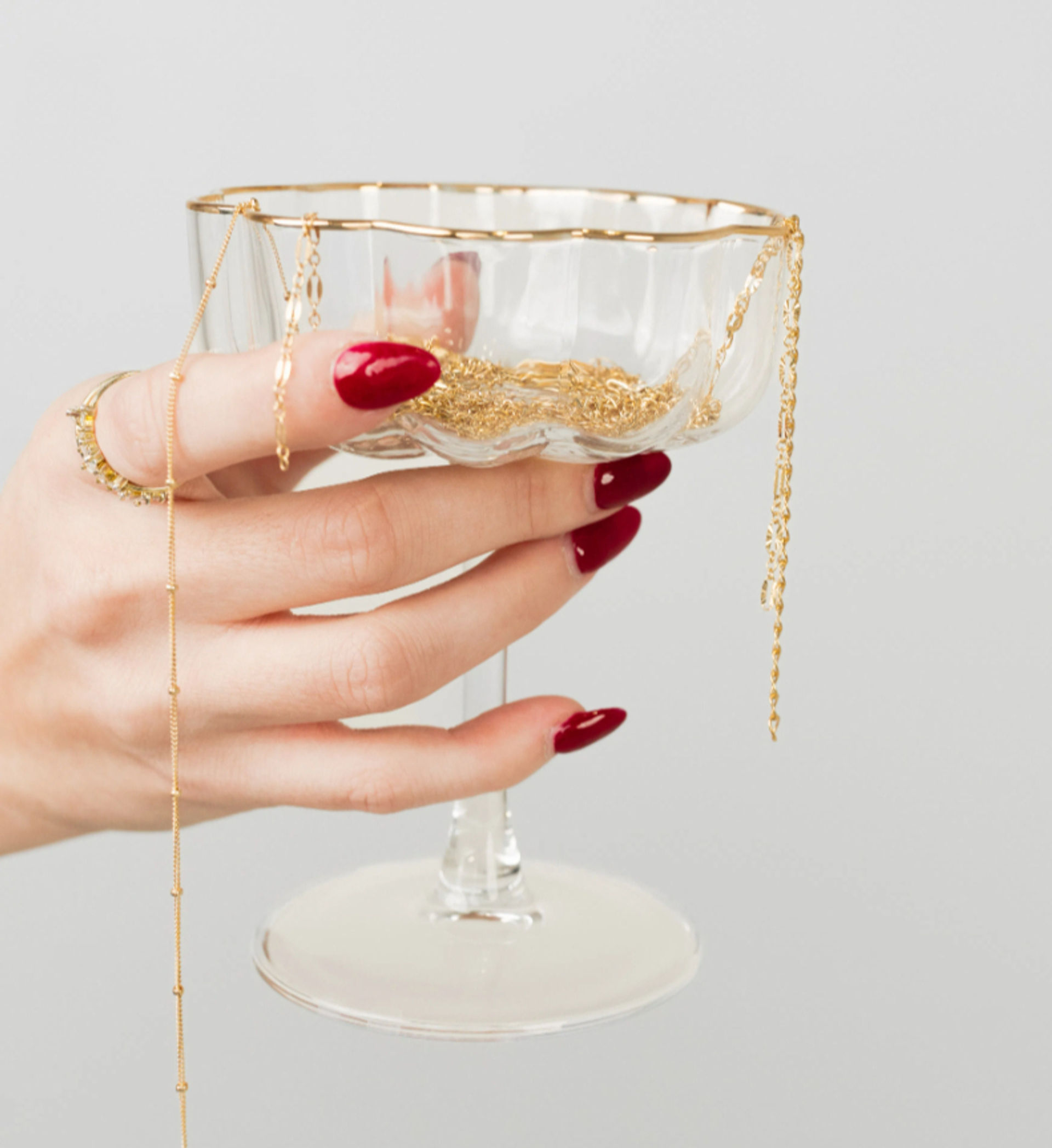 Close-up jewelry display: hand with glossy red manicure and gold rings holds a glass coupe filled with delicate gold chains and necklaces against a light gray background.