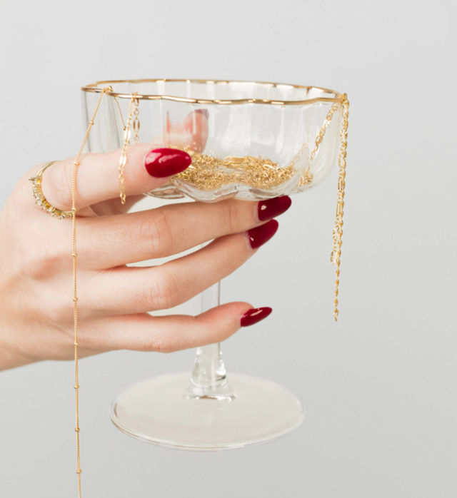 Close-up jewelry display: hand with glossy red manicure and gold rings holds a glass coupe filled with delicate gold chains and necklaces against a light gray background.