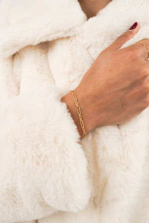 Close-up of wrist wearing a delicate gold paperclip bracelet and thin ring with burgundy nails on a cream faux-fur coat — cozy luxe fashion accessory