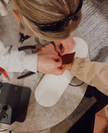 Top-down view of a nail technician applying a gold nail charm to a client's press-on nail on a white hand rest, with pliers and tools on the table