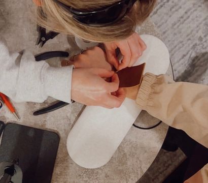 Top-down view of a nail technician applying a gold nail charm to a client's press-on nail on a white hand rest, with pliers and tools on the table