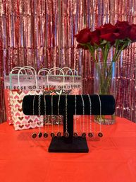 Valentine's Day retail display with numbered charm bracelets on a black velvet stand, bouquet of red roses in a vase, heart-patterned gift bags and shimmering rose-gold fringe backdrop on a red table.